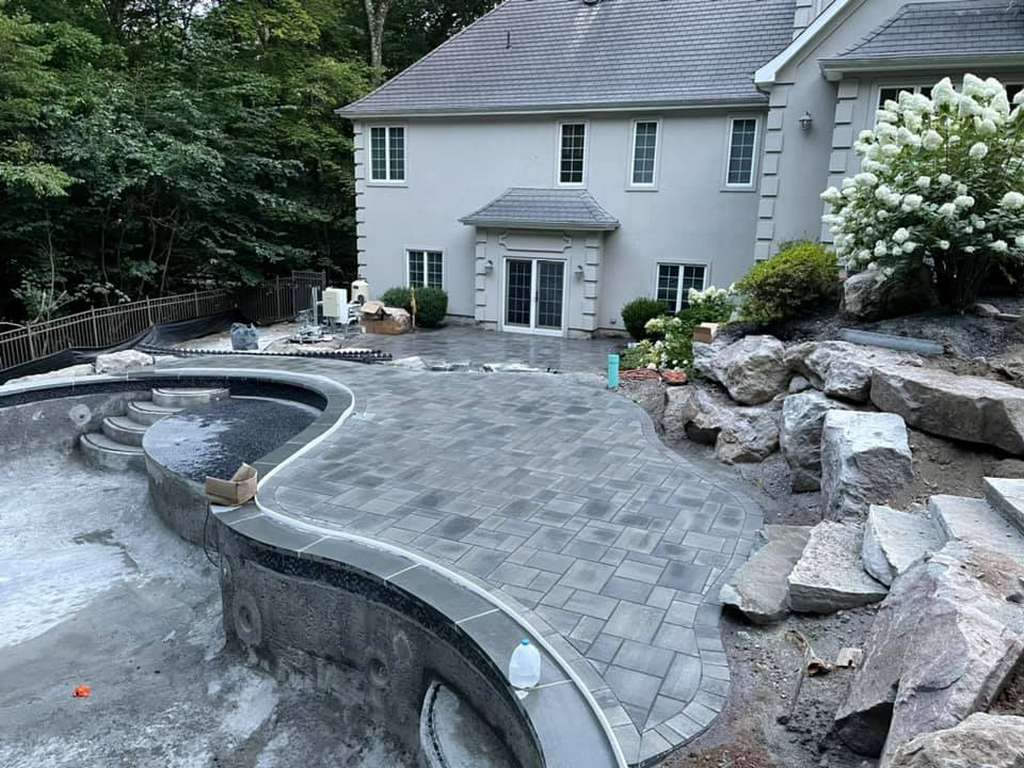 Custom paver pool patio with bluestone coping and natural boulder retaining wall behind a New Jersey home